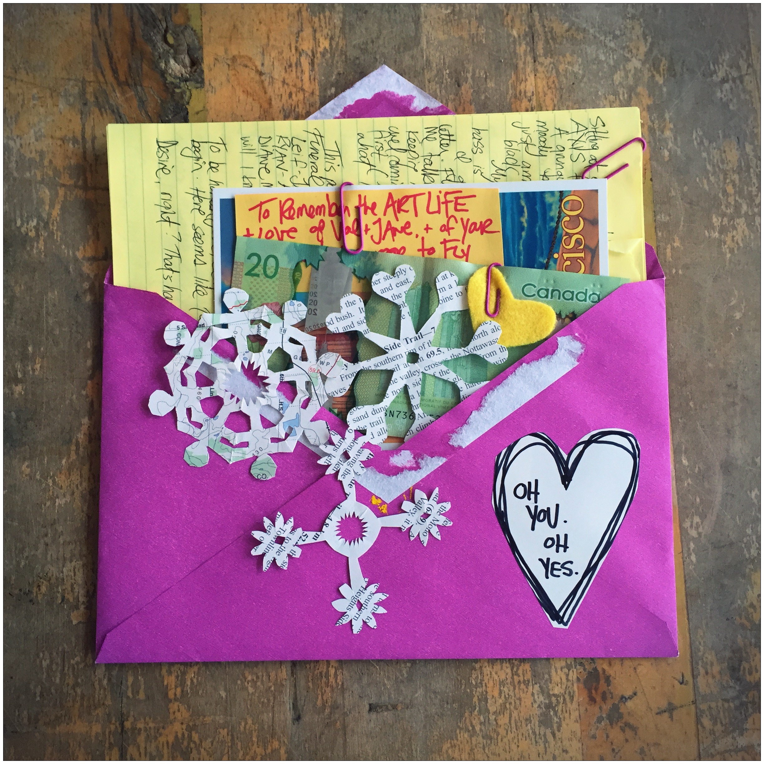 A purple envelope filled with a birthday letter, written in black ink on a yellow sheet of lined paper. There are some homemade snowflake cut-outs in the foreground as well as a heart cut-out that reads, "Oh you. Oh yes." This is by Danette Relic. 
