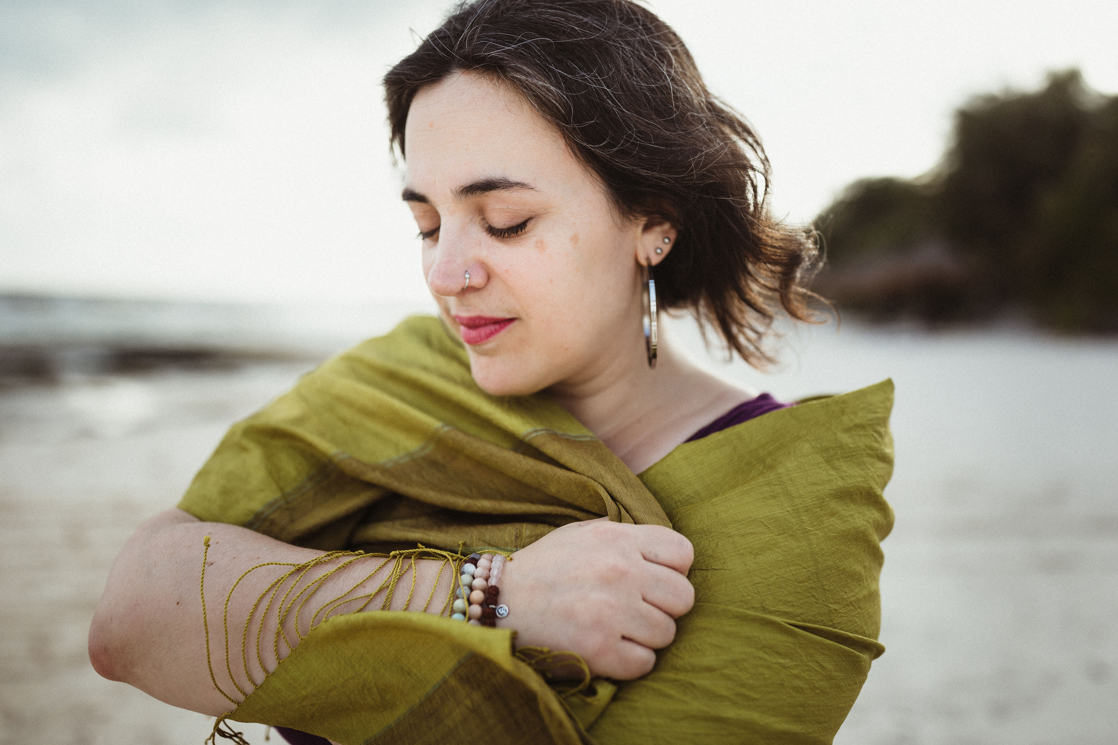 This is a close-up shot of Danette Relic standing on a beach wearing a green shall, a purple dress, three beaded bracelets and three different types of earrings - one a big hoop and two unique studs. Her hair is wavy and blowing in the wind. This is by Shannon Laliberte.