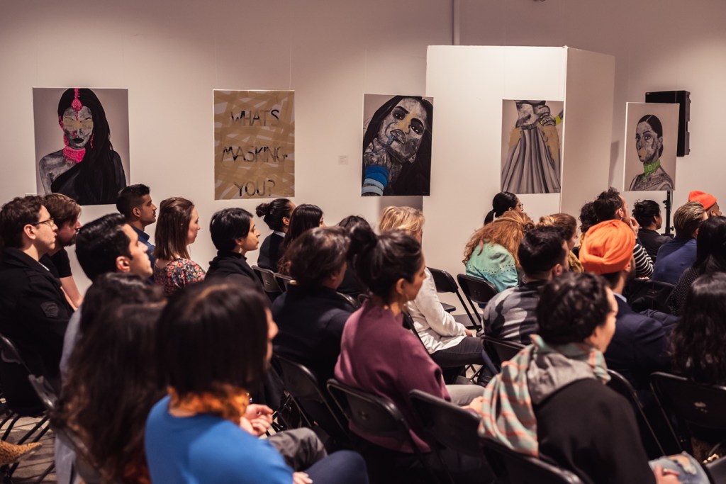 The audience at Romana Kassam's MasksxMolds exhibiition are seated during a presentation. Romana's works are hung in a gallery throughout the room, alongside a print that reads, "What's masking you?" Photo by: Nabeel Pervaiz.