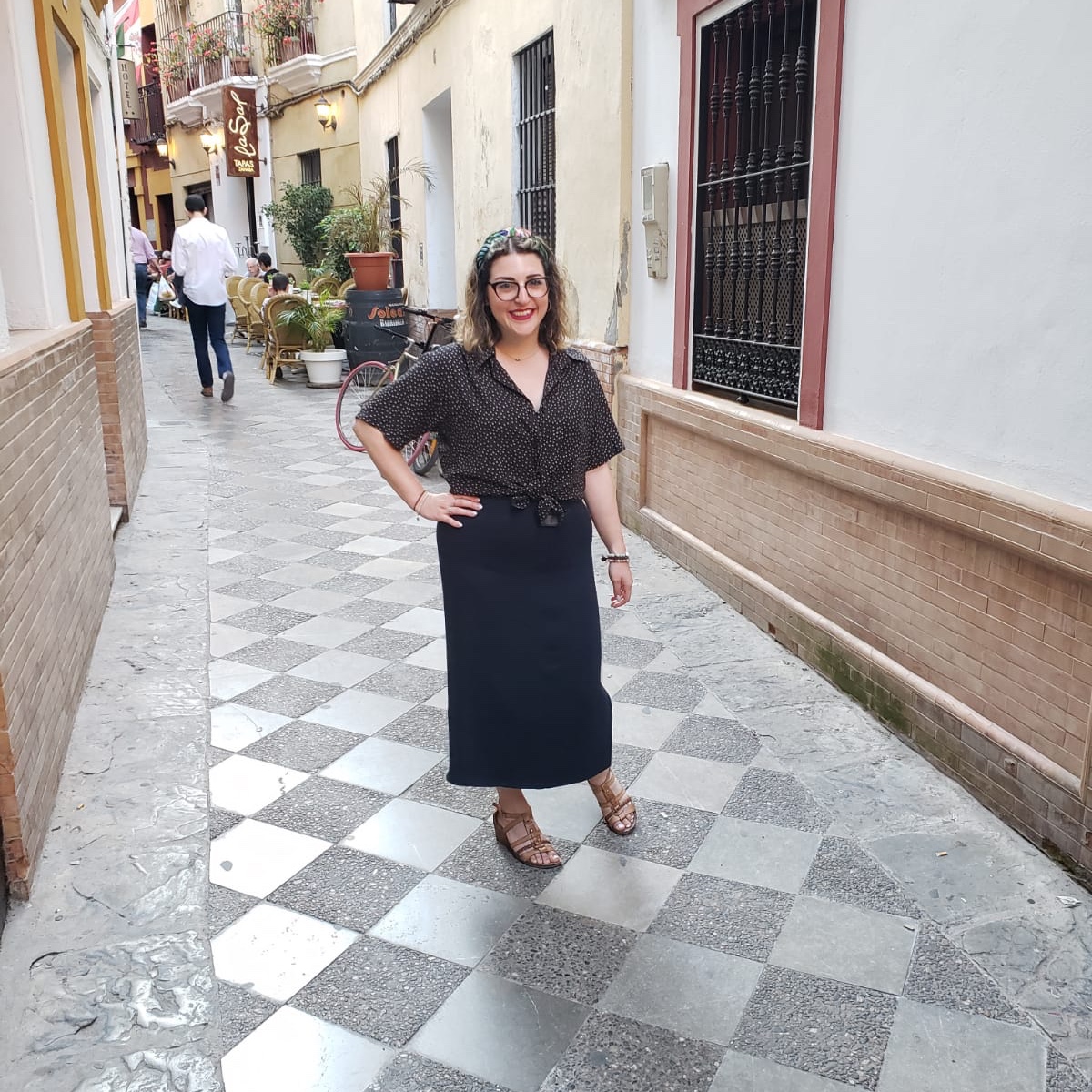 Author Leviana Coccia stands in an alleyway in Sevilla, Spain. She is wearing brown wedged sandals, a long blue skirt, a polka dot blouse and a colourful headband. Photo courtesy of: Leviana Coccia.