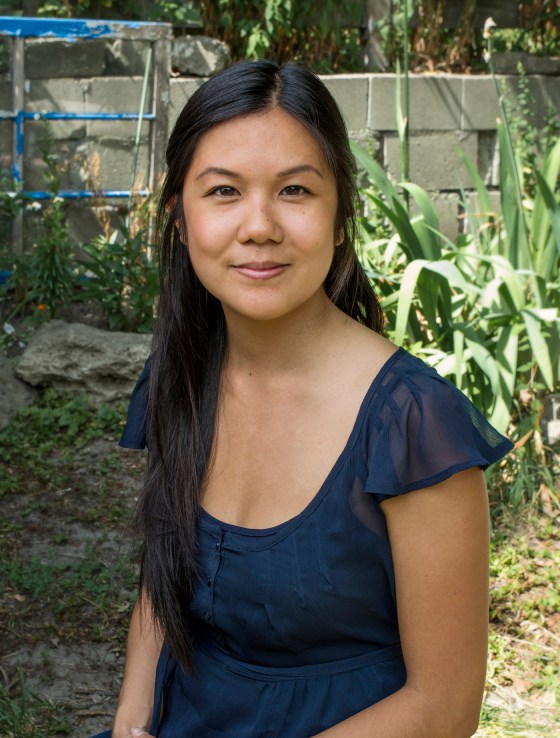 Andrea Kwan is a Registered Social Worker, Psychotherapist. In the image, Andrea is sitting in a green space, in the sunshine, wearing a navy blue blouse. Her hair is dark, straight, long and pulled to one side. This photo is by: Davide Burzotta.